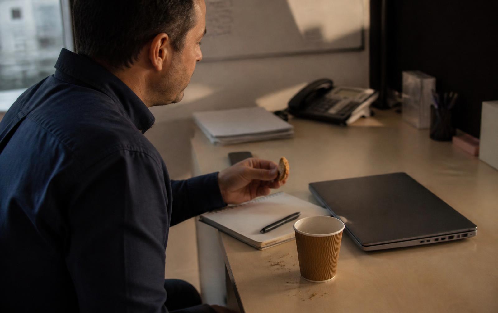 Dans un bureau ordinaire en fin de journée, un adulte vu de dos prend un biscuit à côté d'un gobelet de café, d'un carnet ouvert et d'un ordinateur portable fermé.