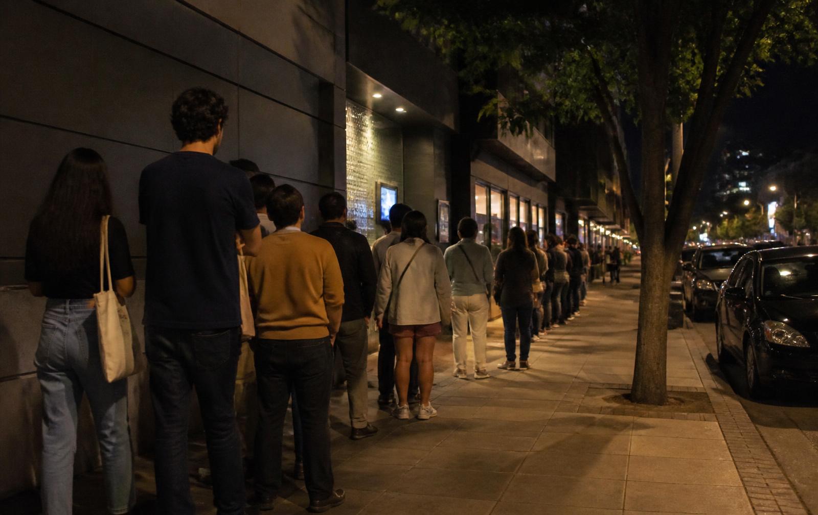 Sur un trottoir d'une autre grande ville, la nuit, une file de spectateurs attend le long de la façade sombre d'un cinéma, de dos, sous l'éclairage ordinaire de la rue.