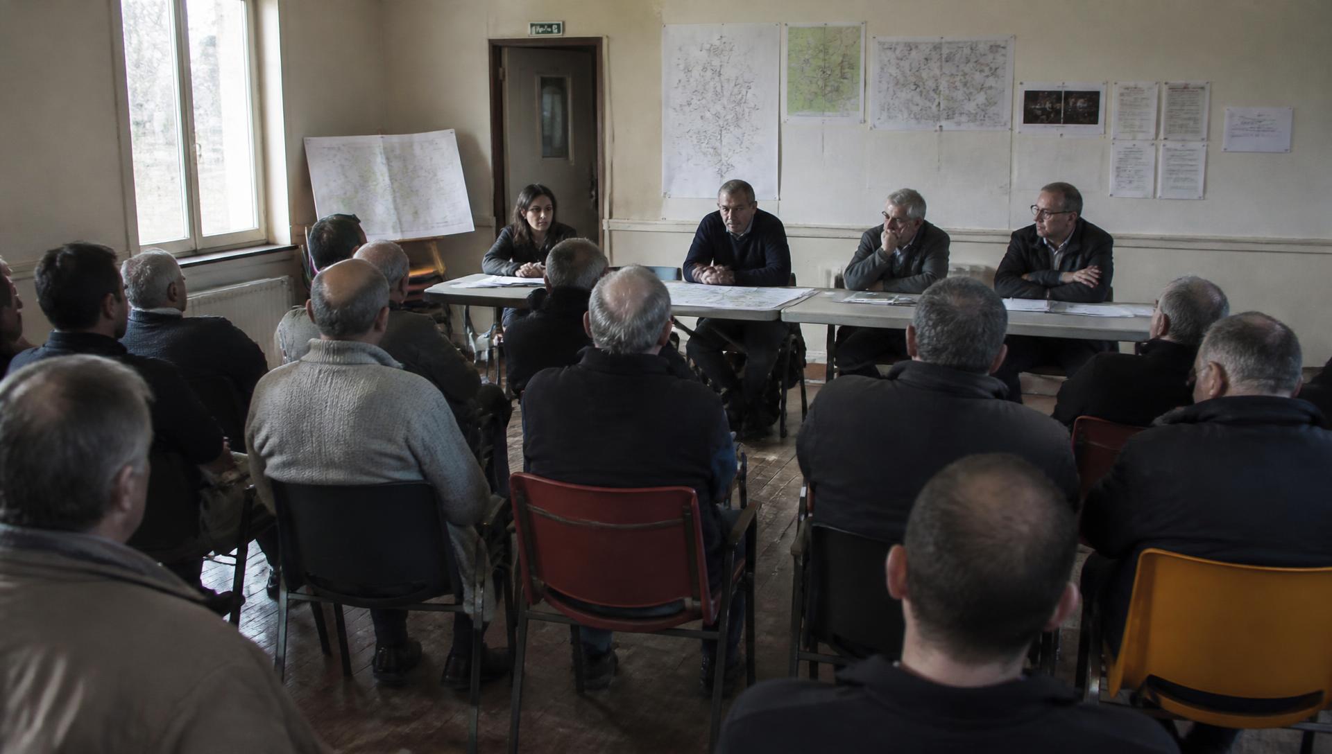 Réunion publique dans une salle communale rurale, habitants assis face à une table de présentation et des cartes affichées.