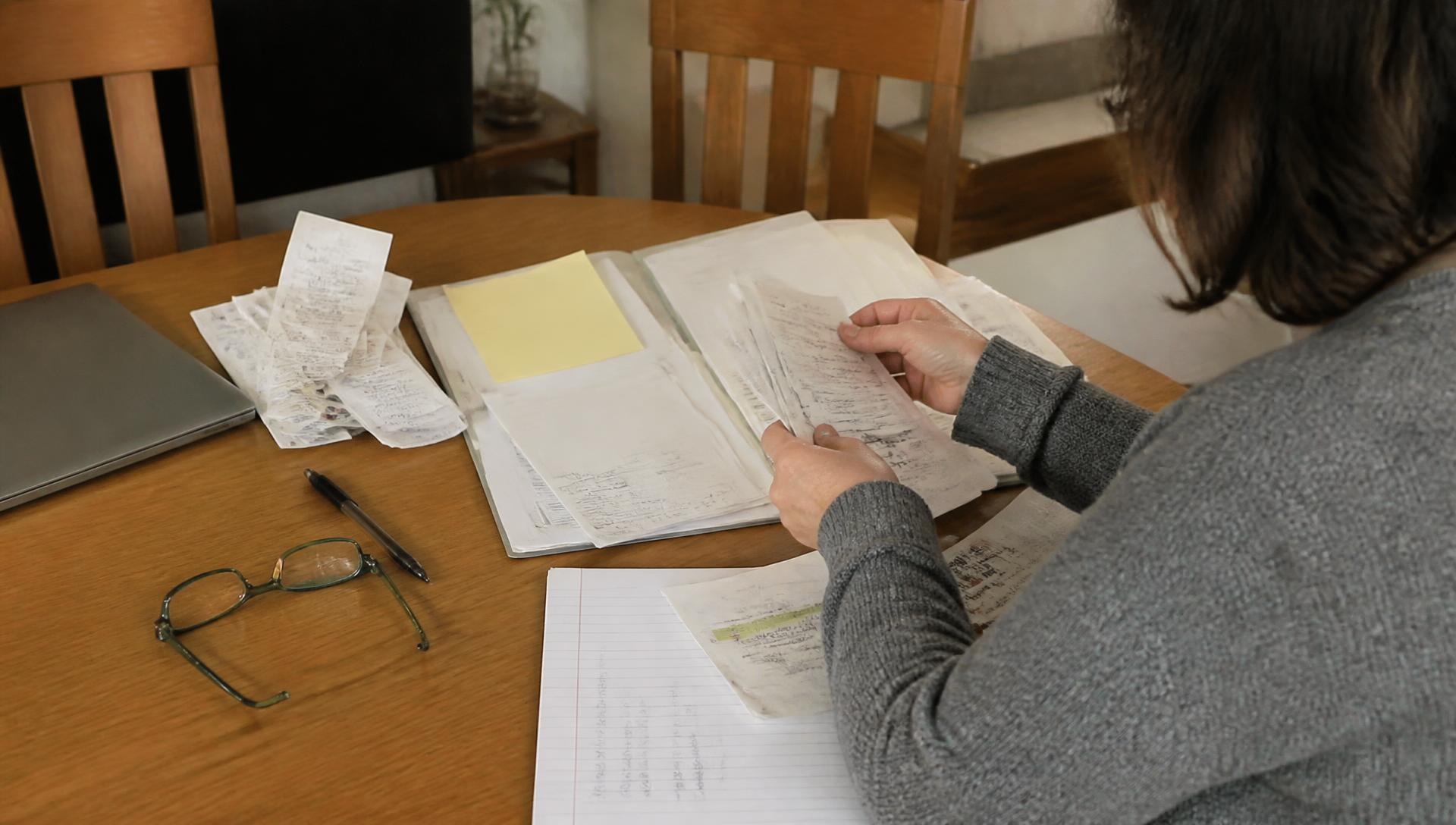 À une table de salle à manger, une assurée trie factures, ordonnances et formulaires dans un classeur pour justifier un dossier de remboursement.