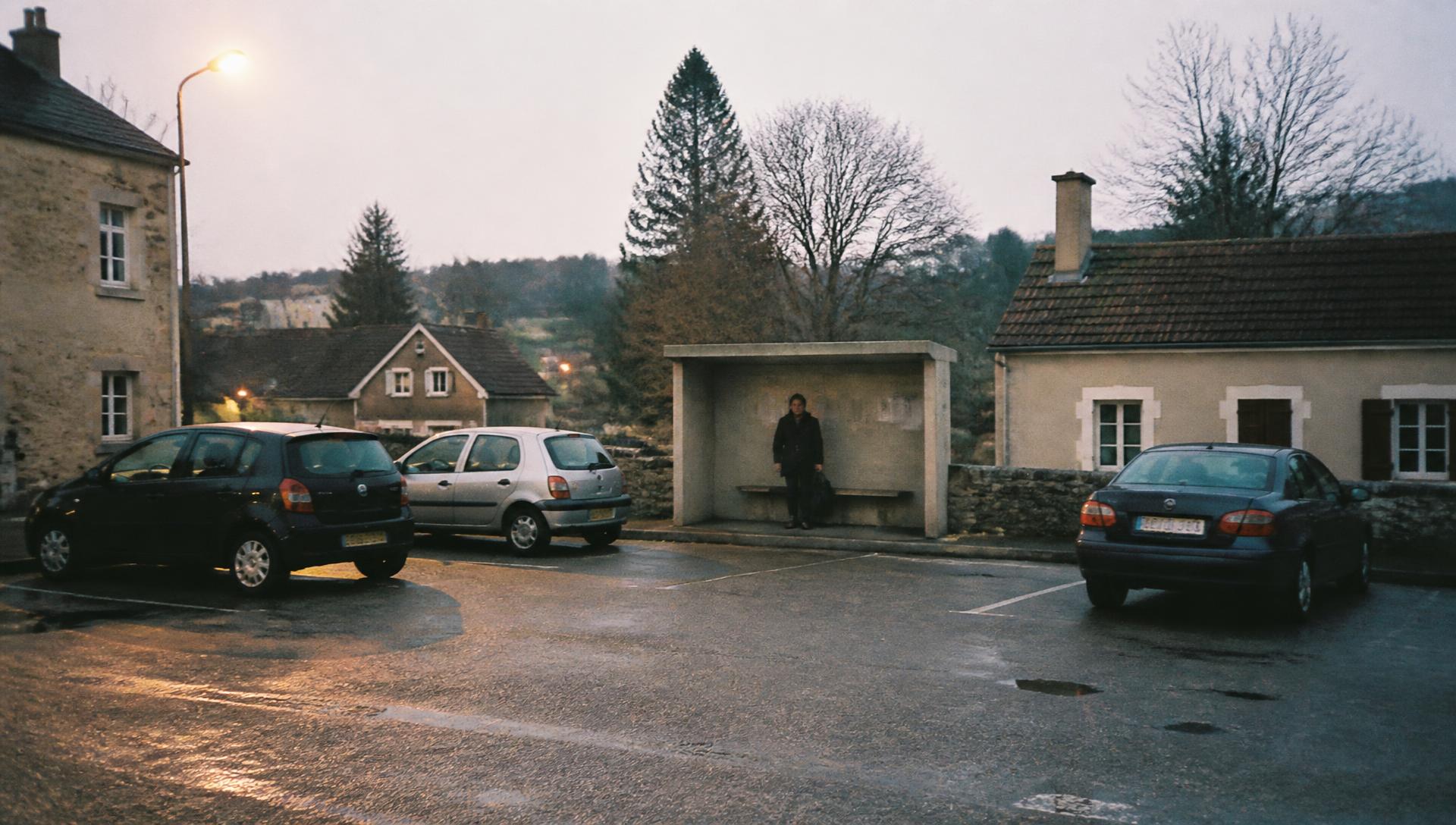 Parking d'un village rural au crépuscule, quelques voitures garées et une personne seule attendant sous un abri.