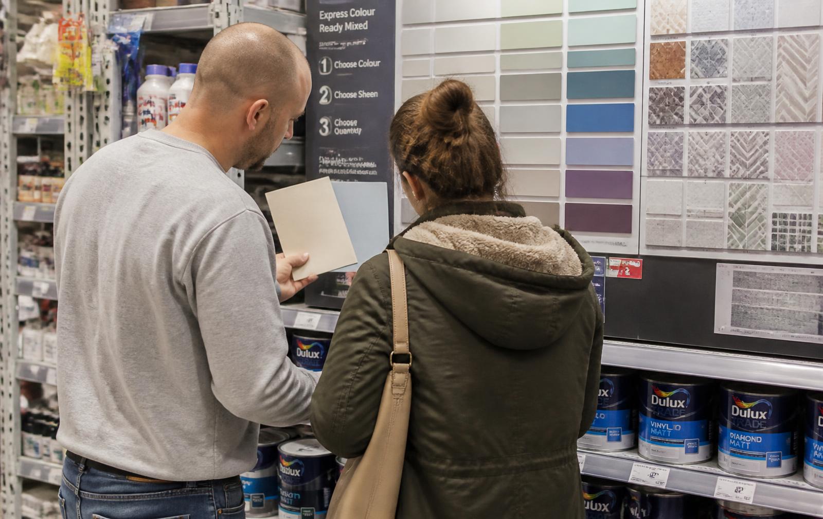 Dans un rayon peinture d'un magasin de bricolage, deux clients comparent des échantillons de couleurs et de carrelage, de dos, sous la lumière ordinaire du magasin.