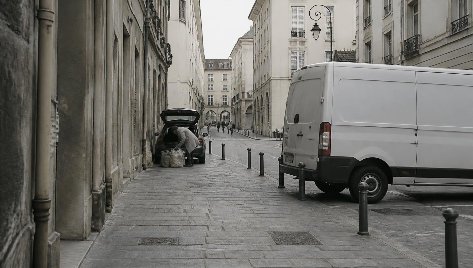 Bord d'un périmètre piétonnisé : une camionnette de livraison arrêtée près des bornes, une résidente déchargeant des courses volumineuses du coffre de sa voiture.