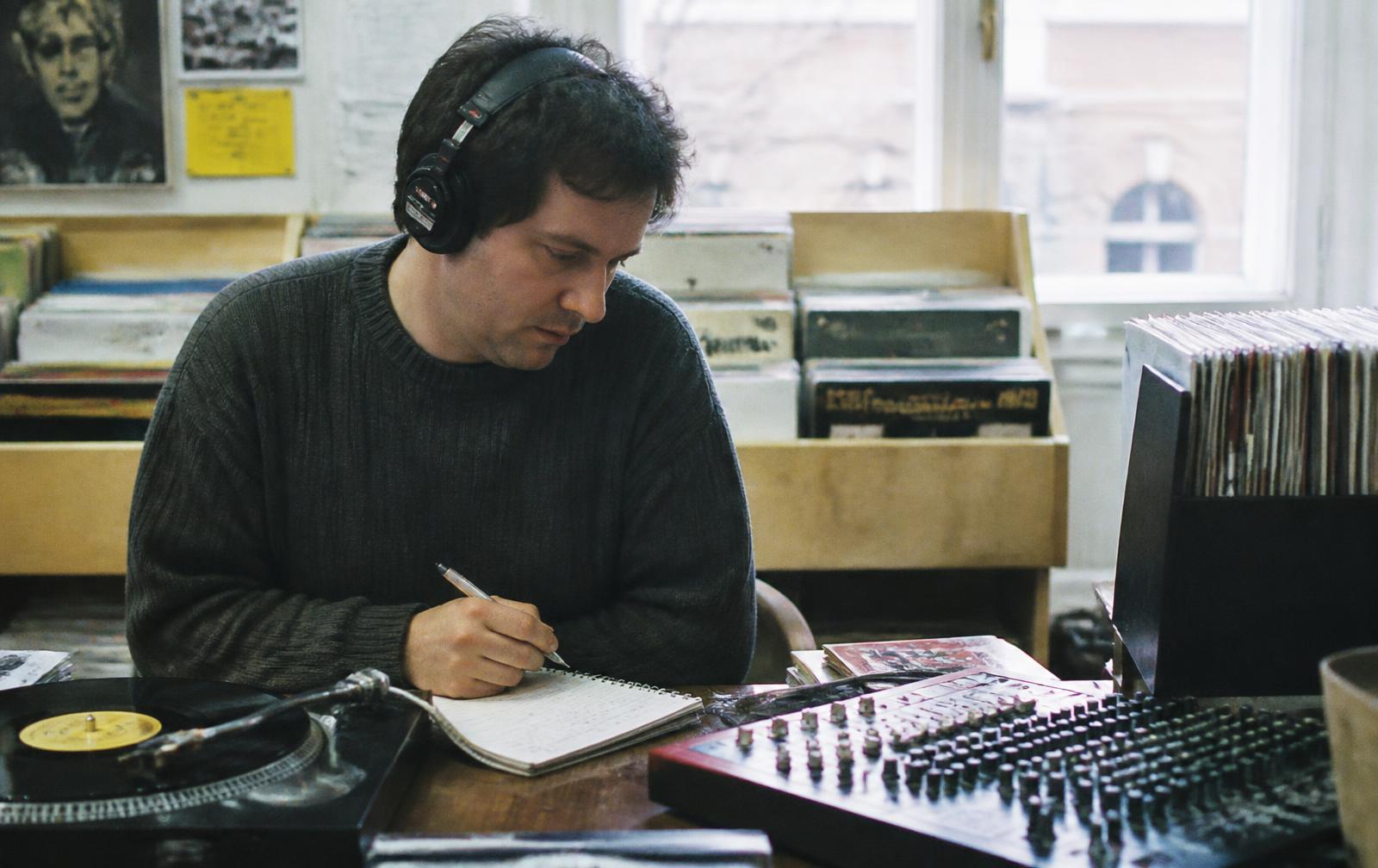 Dans un disquaire indépendant, un journaliste ou programmateur écoute un morceau au casque devant une platine et une petite console, en prenant des notes sur un carnet.