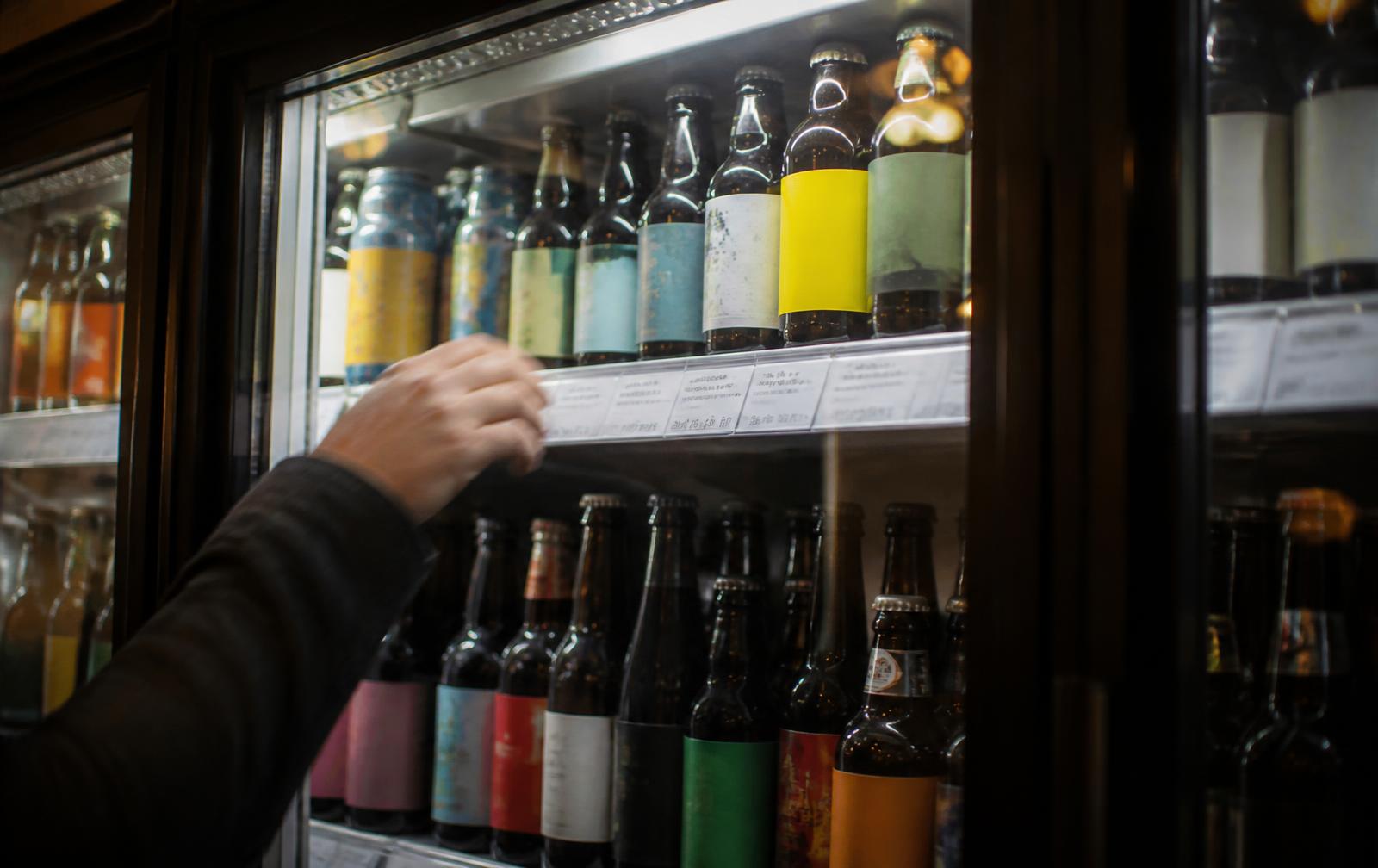 Frigo vitré d'un bar rempli de bouteilles et canettes de couleurs et de formes variées, étiquettes illisibles ; une main floue se tend vers l'une des bouteilles.