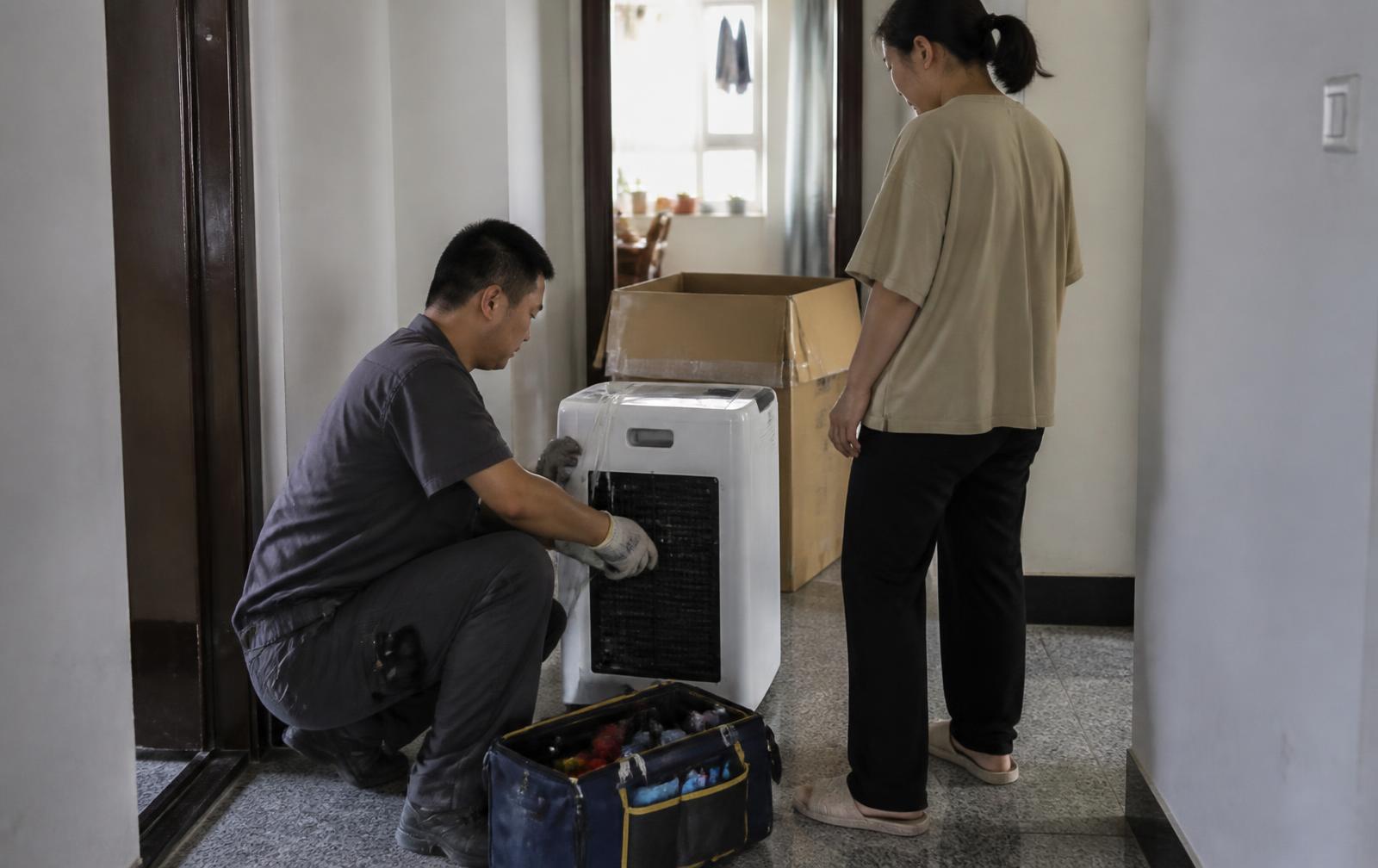Dans le couloir d'un appartement ordinaire, un technicien accroupi intervient sur un appareil électroménager à côté d'un carton d'emballage, sous le regard de l'habitante vue de dos.