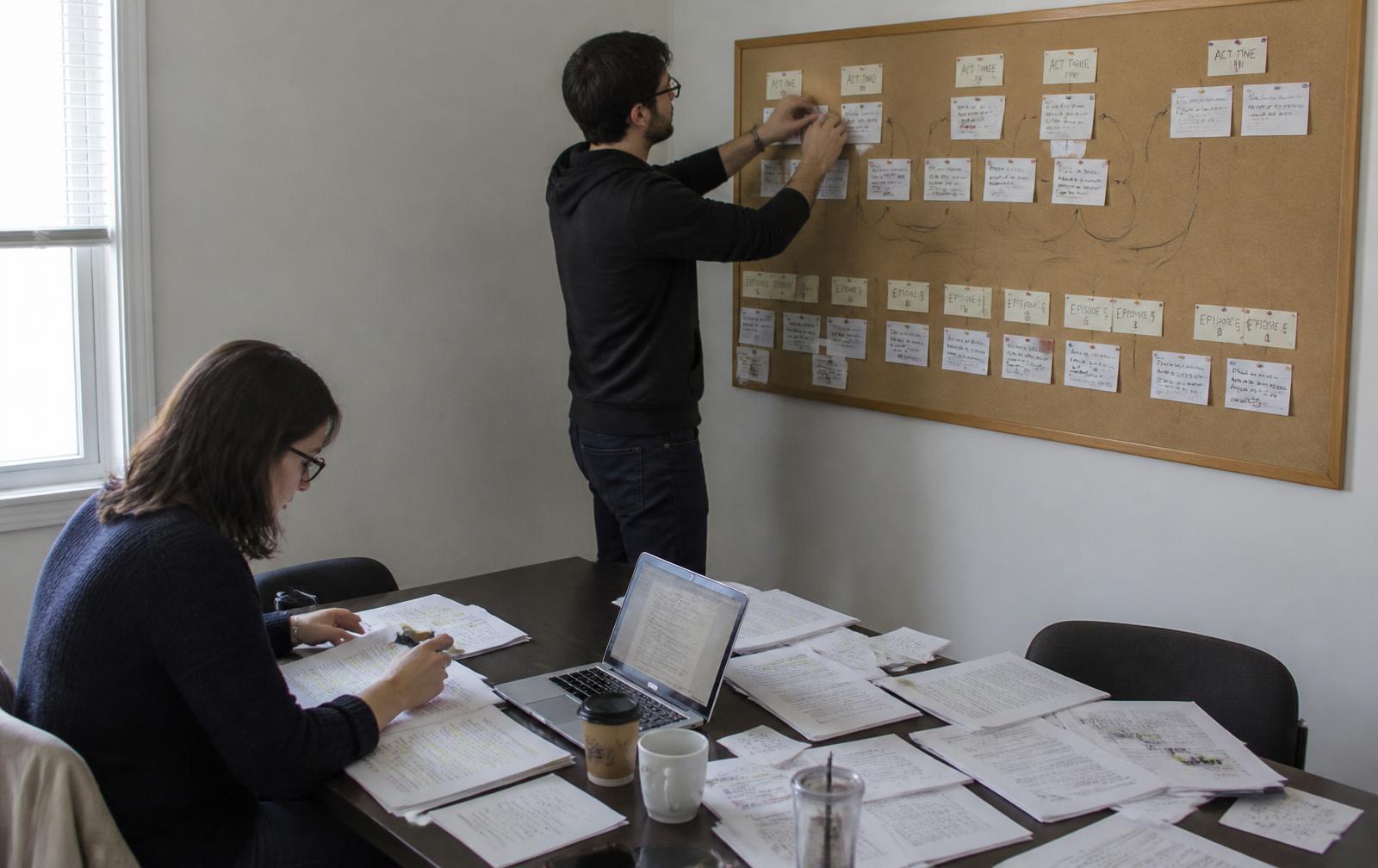 Deux auteurs dans une petite salle de travail : pages de scénario annotées sur la table, ordinateur portable ouvert, tableau de liège couvert de fiches manuscrites reliées par des flèches d'arcs narratifs, l'un des deux déplaçant une fiche.