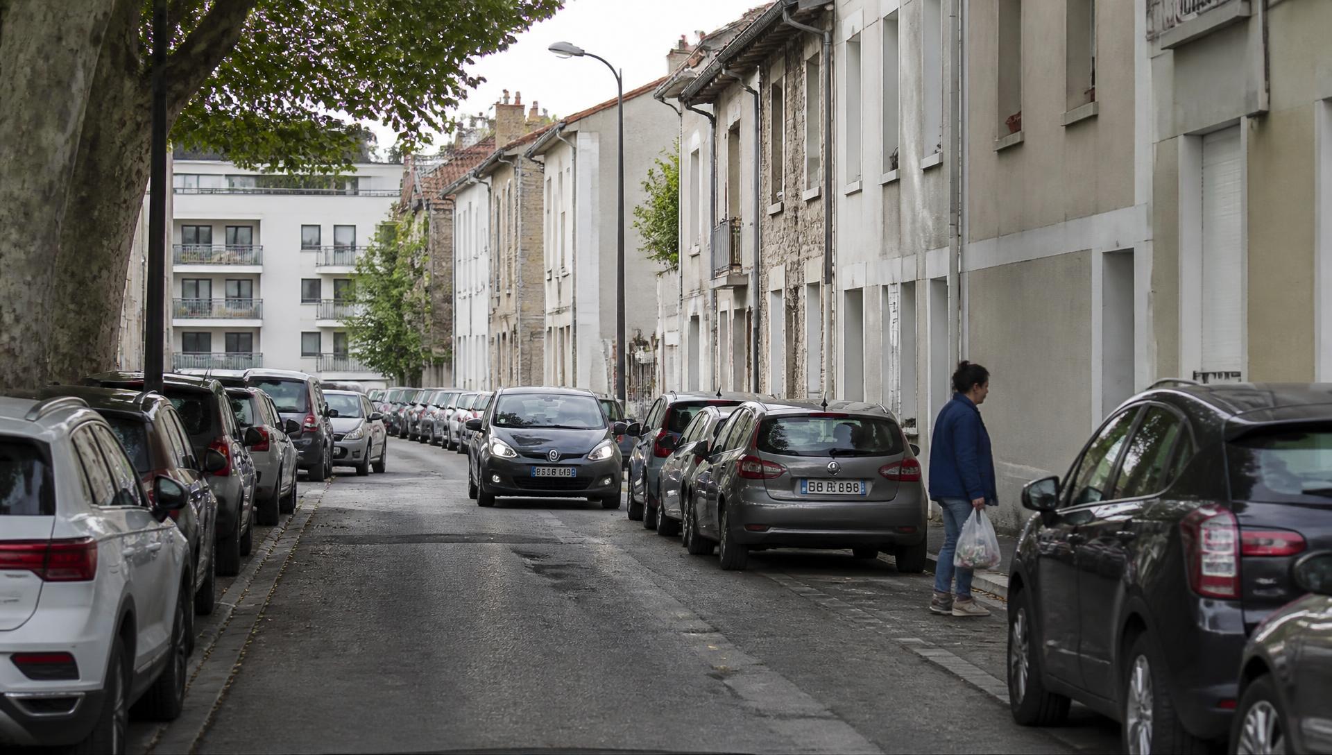 Rue résidentielle proche du centre : voitures garées pare-chocs contre pare-chocs des deux côtés, une automobiliste cherchant une place, une habitante rentrant avec ses courses.
