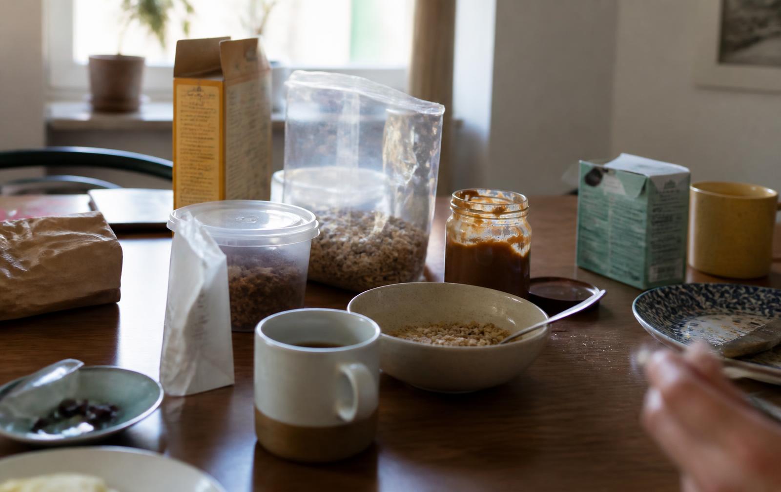 Table de petit-déjeuner ordinaire dans une cuisine : plusieurs paquets et bocaux ouverts, un bol de céréales, une tasse de café, une boîte de thé, vaisselle dépareillée, aucune marque identifiable.