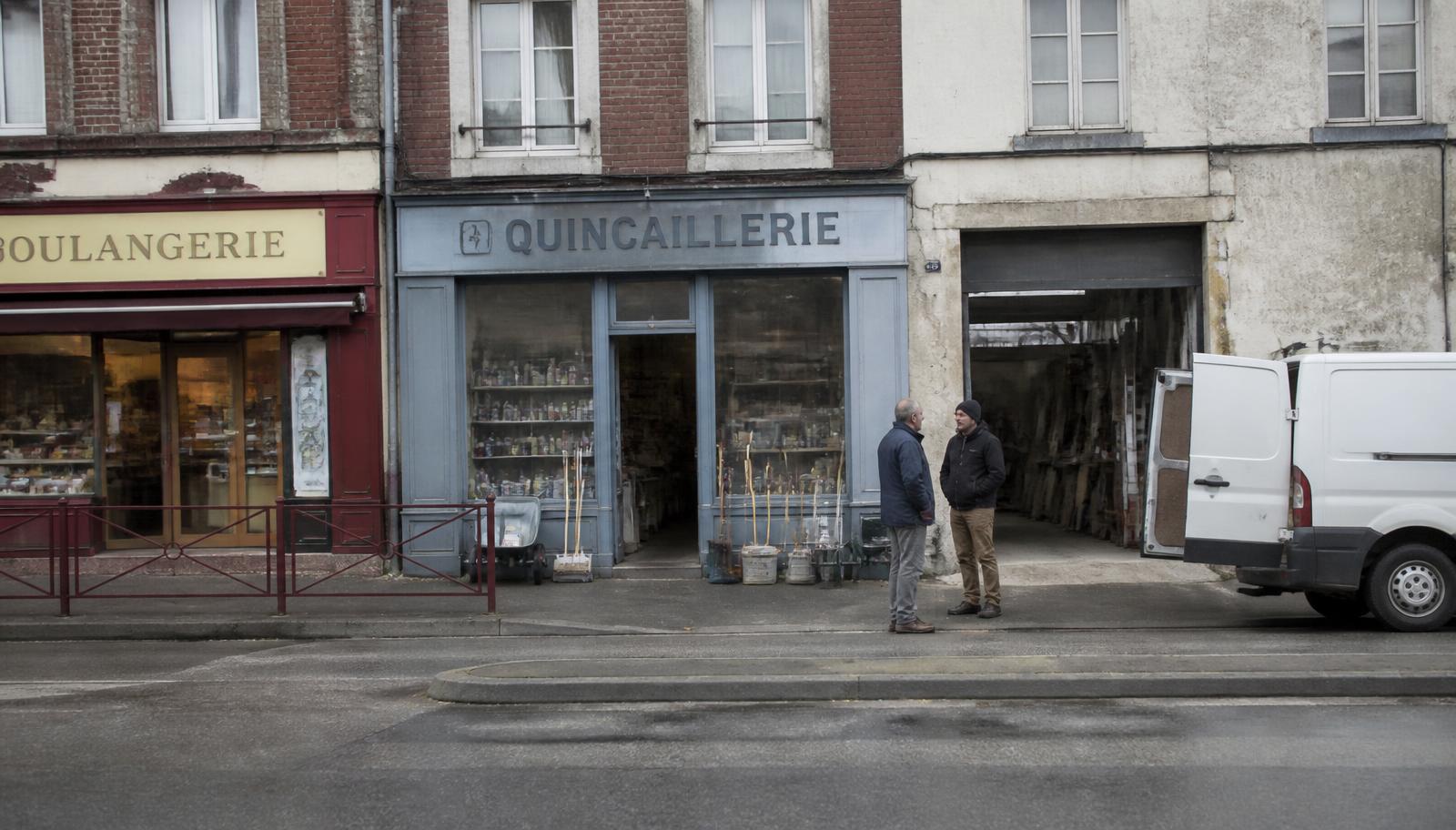 Rue commerçante d'un bourg industriel : une boulangerie, une quincaillerie, l'entrée d'un atelier et une camionnette de livraison ; deux hommes discutent sur le trottoir.