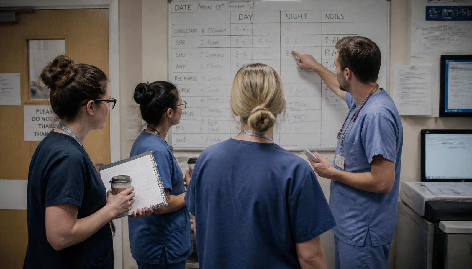Transmission courte entre soignants devant un tableau de service manuscrit dans un local hospitalier : quatre professionnels en tenue debout, l'un désignant une ligne du tableau, les autres écoutant, carnets et gobelets à la main.