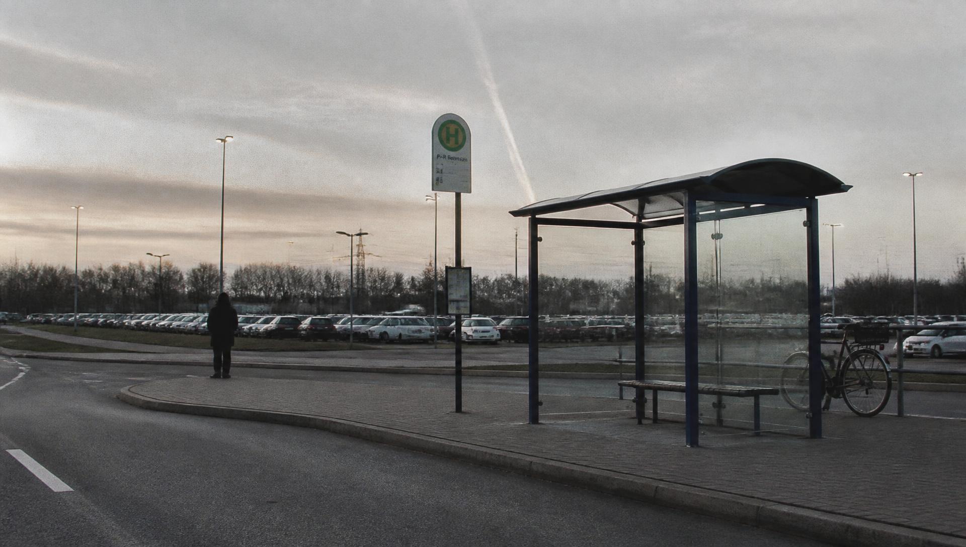 Au petit matin, un abri de bus et un parking relais périphérique aux trois quarts vides, une silhouette isolée en attente.
